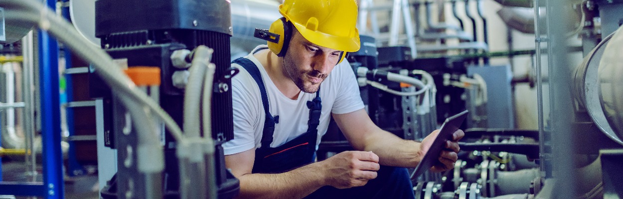 Focused plant worker in overalls, with protective helmet on head and antiphons on ears using tablet for checking machine while crouching.